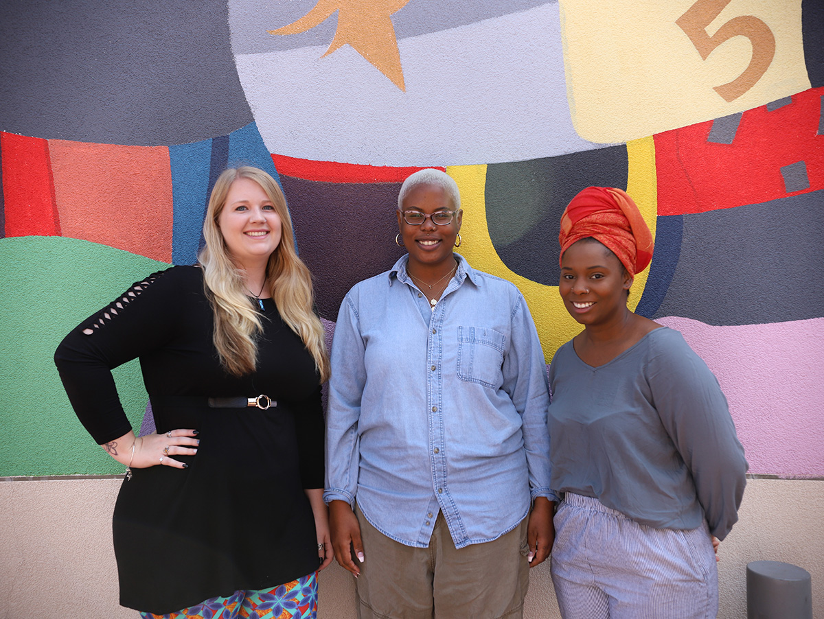 Photo of three woman smiling and posing in front of a colorful mural