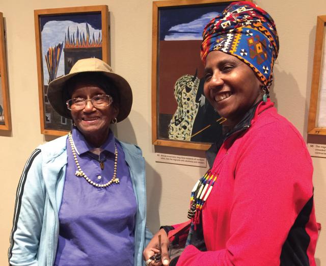 Photograph of two older women smiling in front of panels from Jacob Lawrence's Migration Series.