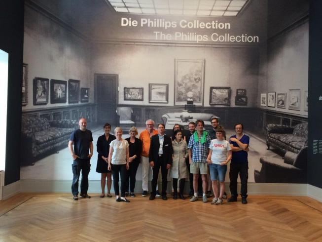 Photograph of a group of people in a big room, posing in front of a wall with a reproduction of an archival photograph of The Phillips Collection