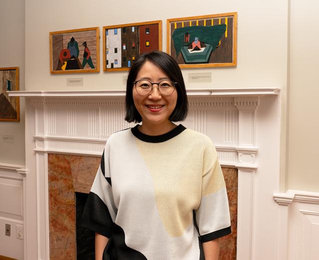 Headshot of Tie Jojima with Jacob Lawrence's Migration Series on the wall behind her