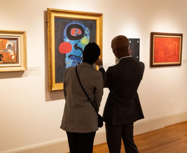 Two people looking at 4 paintings on a wall in a gallery