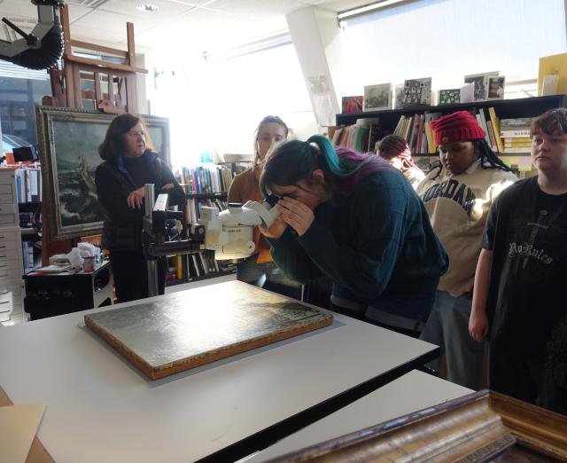 A high school student looks at a painting through a microscope with other students around her