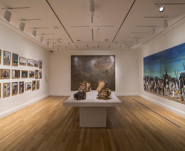 Installation view of The Warmth of Other Suns: Stories of Global Displacement. On the walls left to right: Jacob Lawrence’s Migration Series (1940-41), Nari Ward’s Breathing Panel, Oriented Right (2015), and Benny Andrews, Trail of Tears (2005). In the center: Beverly Buchanan sculptures