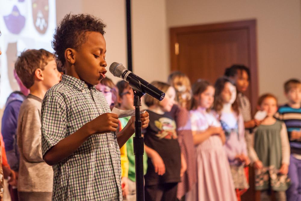 Photograph of elementary school students on stage, with one boy at a microphone reading off a paper