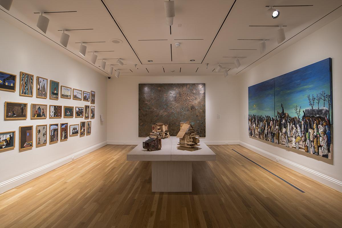 Installation view of The Warmth of Other Suns: Stories of Global Displacement. On the walls left to right: Jacob Lawrence’s Migration Series (1940-41), Nari Ward’s Breathing Panel, Oriented Right (2015), and Benny Andrews, Trail of Tears (2005). In the center: Beverly Buchanan sculptures