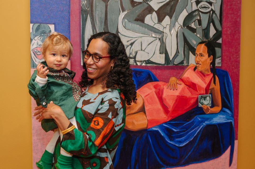 Photograph of Mequitta Ahuja holding her son in front of her painting Xpect