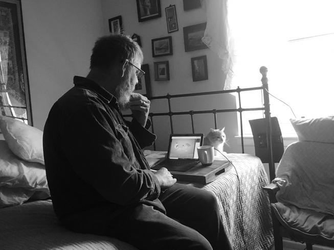 Black and white photograph of a person sitting on a bed with a computer and a cat