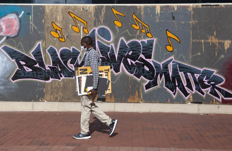 Photograph of a man walking in front of a Black Lives Matter mural, holding a sign