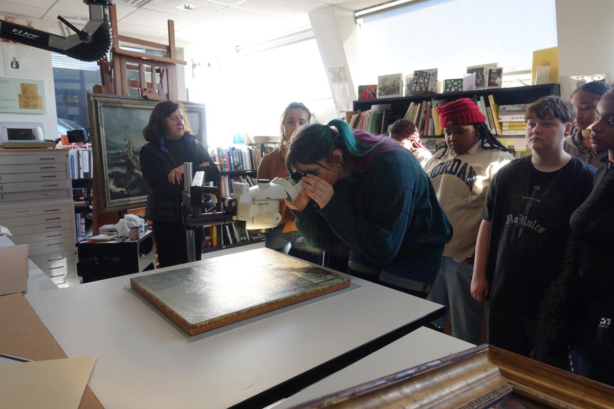 A high school student looks at a painting through a microscope with other students around her